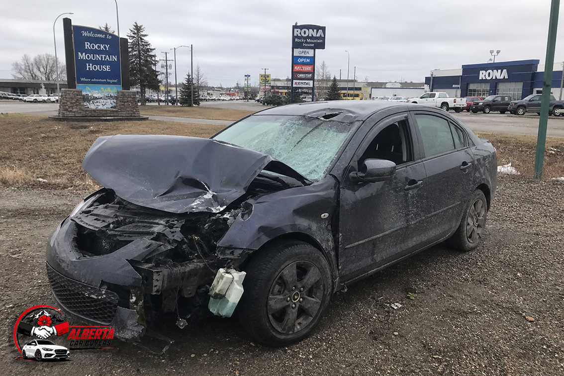 Wrecked dark sedan with severe front-end damage parked on a gravel lot near a “Welcome to Rocky Mountain House” sign and a RONA store under an overcast sky.