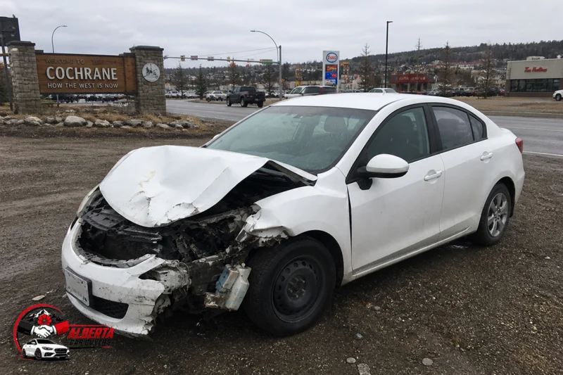 White sedan with heavy front-end damage parked on a muddy roadside near a “Welcome to Cochrane” sign, with traffic lights, shops, and an overcast sky in the background.