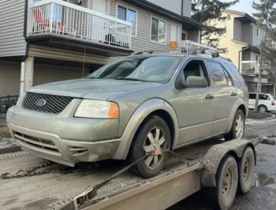 Silver Ford SUV strapped to a trailer in front of beige townhomes with tall evergreen trees under a clear sky.